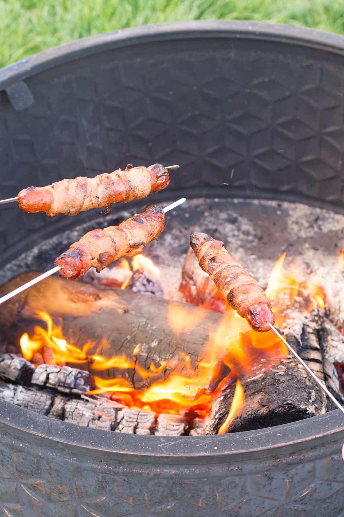 A fire pit with 3 metal skewers roasting bacon wrapped hot dogs!
