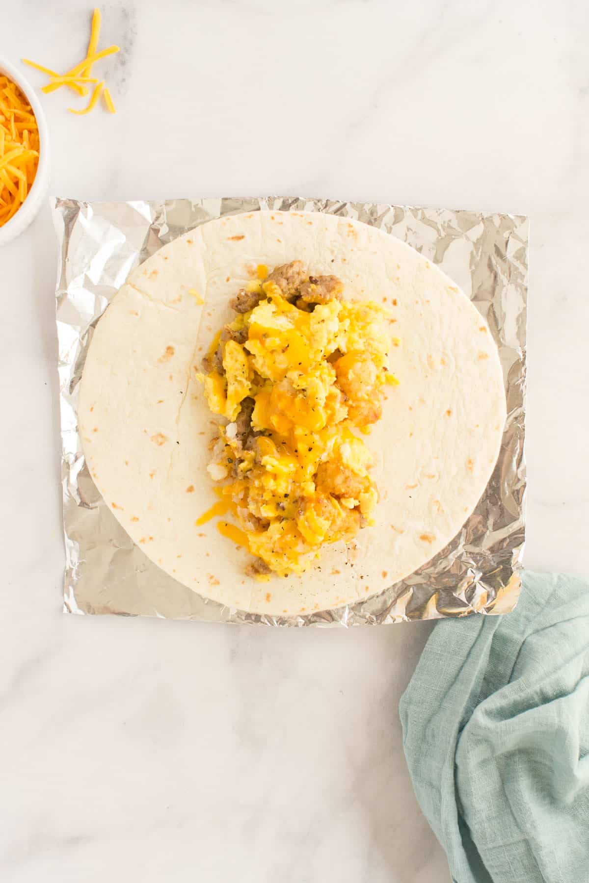 An open breakfast burrito on top of a foil wrapper with a small bowl of shredded cheese on the side.