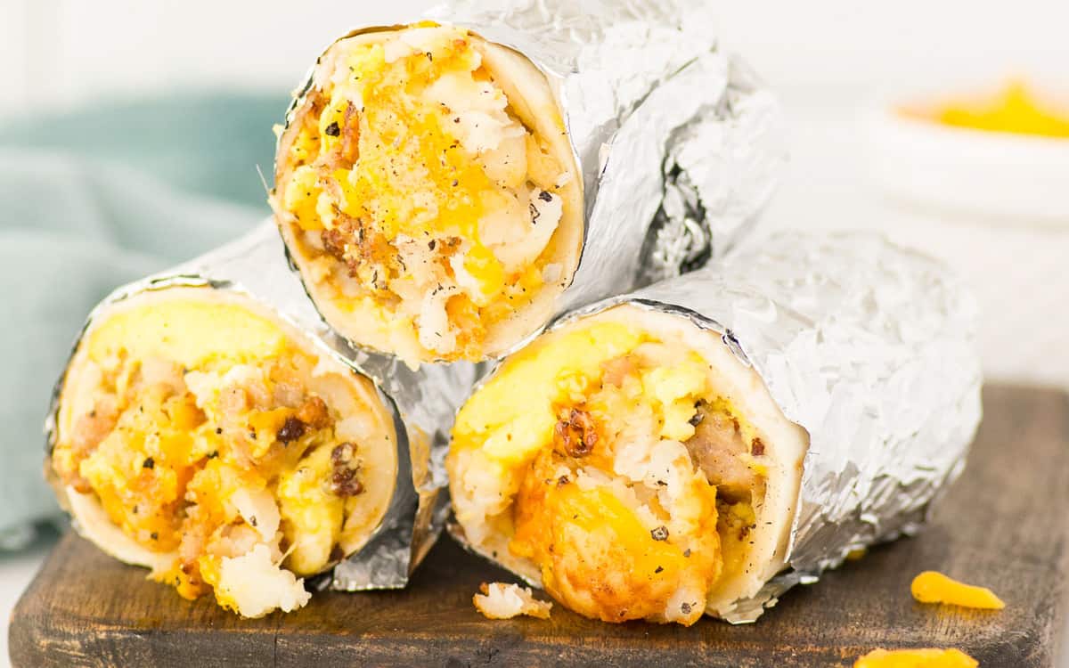 3 owinięte folią burritos śniadaniowe ułożone jedna na drugiej na drewnianej desce.