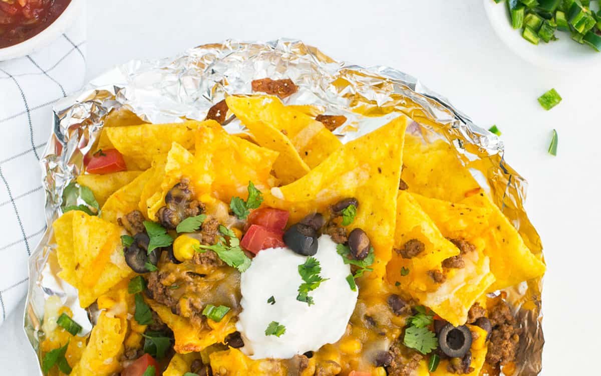 Nachos in a foil packet with a blue and white checked towel with small white bowls full of salsa, jalapenos and cilantro. This is from a list of make ahead camping meals.