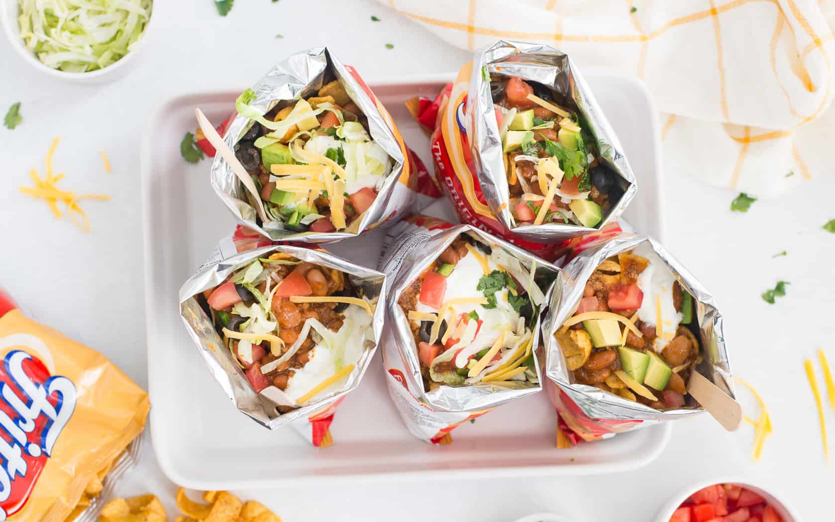 5 bags of walking tacos on a white platter with white bowls filled with lettuce, tomatoes, green onions around it with an open bag of Fritos next to it.