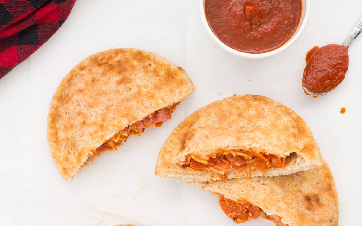 On top of a white plate with a blue rim is holding 5 pita pizza pockets with a small white bowl with cut up pepperoni pieces and a red and black checked towel at the bottom.