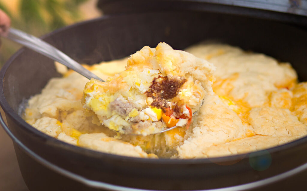 A dutch oven filled with a sausage, eggs and potatoes topped with biscuits.