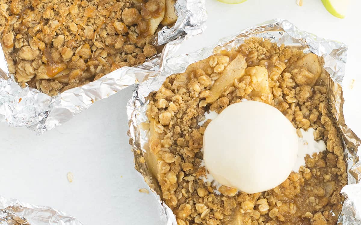 A foil packet filled with warm apples and a cinnamon crumb topping with a scoop of ice cream  on it with 2 other foil packets of warm apples and a cinnamon crumb topping above and below it.  There is a green apple and cinnamon sticks laying beside it too.