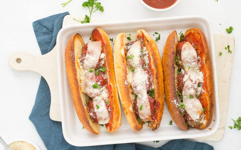 3 meatballs subs next to each other in a white dish on a white cutting board and blue napkin with a bowl of Parmesan cheese in one corner and a bowl of marinara sauce in the other corner.