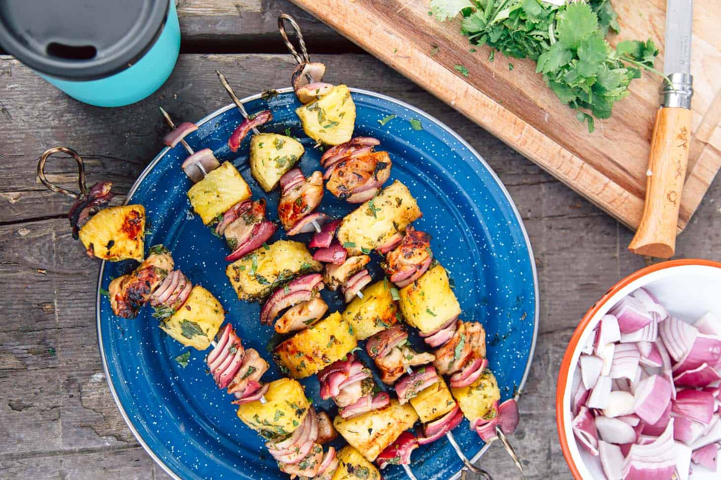 A blue plate with four chicken and pineapple skewers on it with a bowl of onions, herbs and drink on the side.