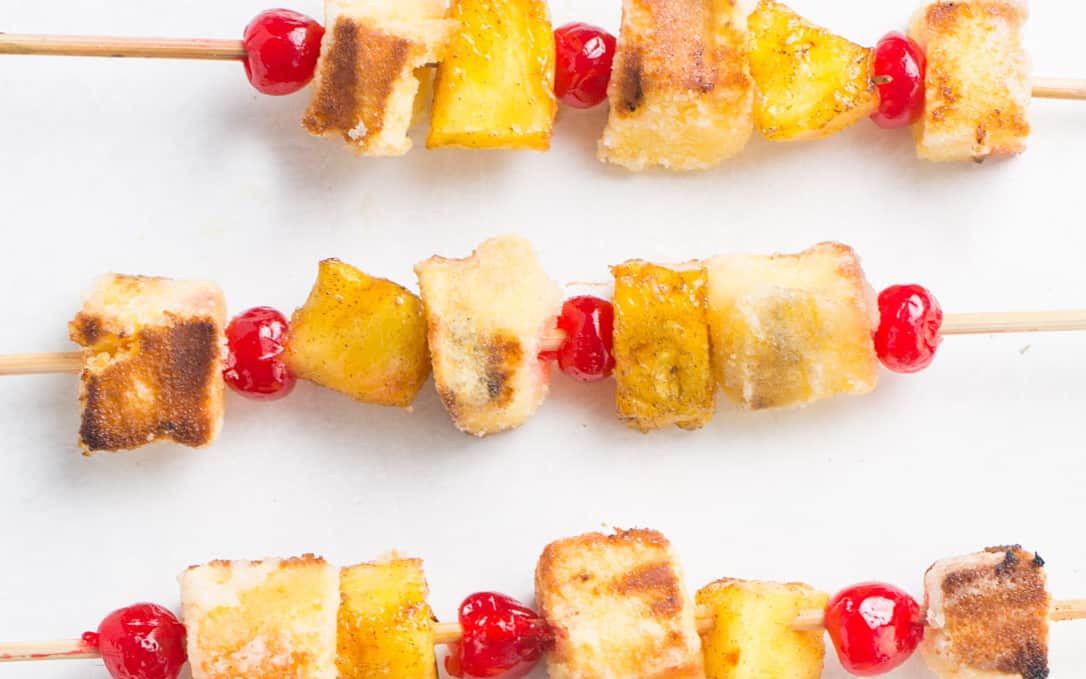 A white oval dish holding 4 pineapple upside down cake kabobs with a striped towel and a pineapple on the side with a small white bowl of cut pineapple on the side. This is from a list of make ahead camping meals.