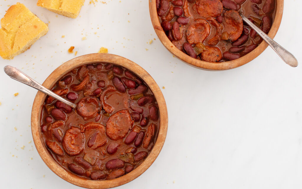 Two wooden bowls of chili with metal spoons in it and a piece of cornbread on the side for a good make ahead camping meals.