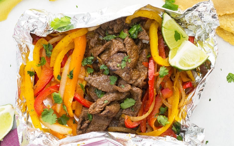 There is a foil packet with fajita toppings under a pink and white towel and cut up limes and tortillas around them.