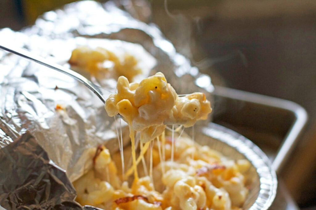 A tinfoil pan filled with noodles and melty cheese with a fork taking a spoonful out of it with strings of cheese down to the pan.
