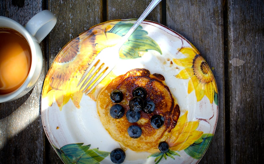 A flowered plate on a picnic table with one pancake with blueberries on it with a fork on the plate and a cup of coffee on the side.