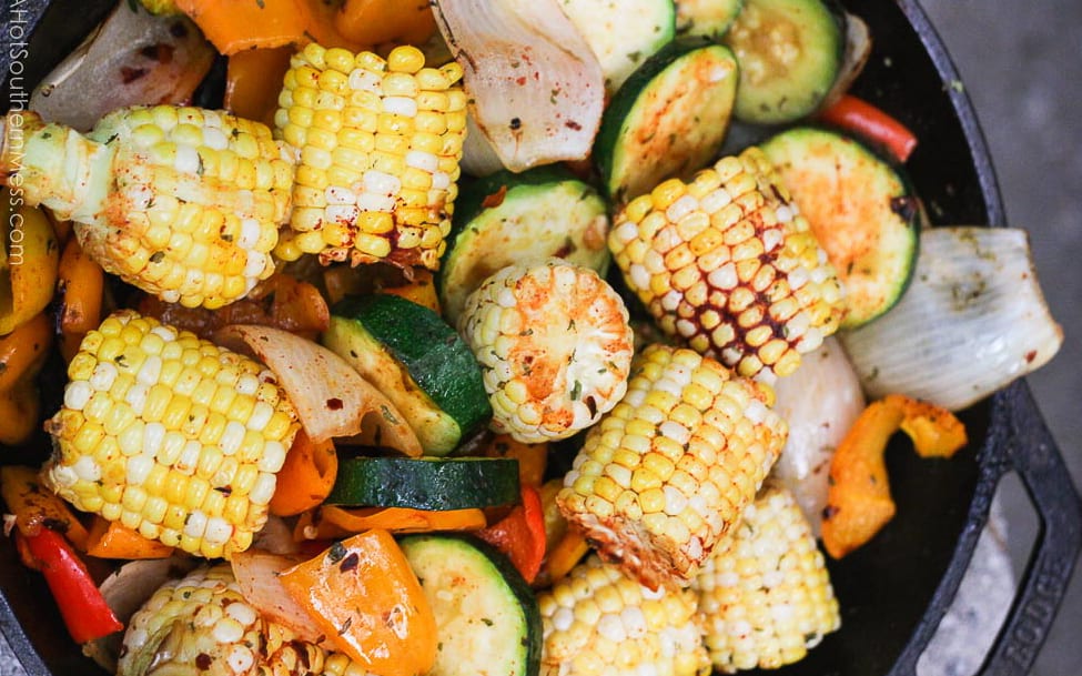 A cast iron skillet with veggies like corn, zucchini, peppers, and onions sprinkled with spices and grilled to perfection for a make ahead camping meal!
