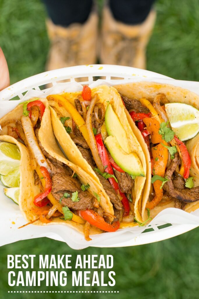 A white plastic basket full of tortillas filled with steak fajitas and limes on the side is being held up with two boots out of focus below. The words Best Make Ahead Camping Meals are in white and underlined in left lower corner.