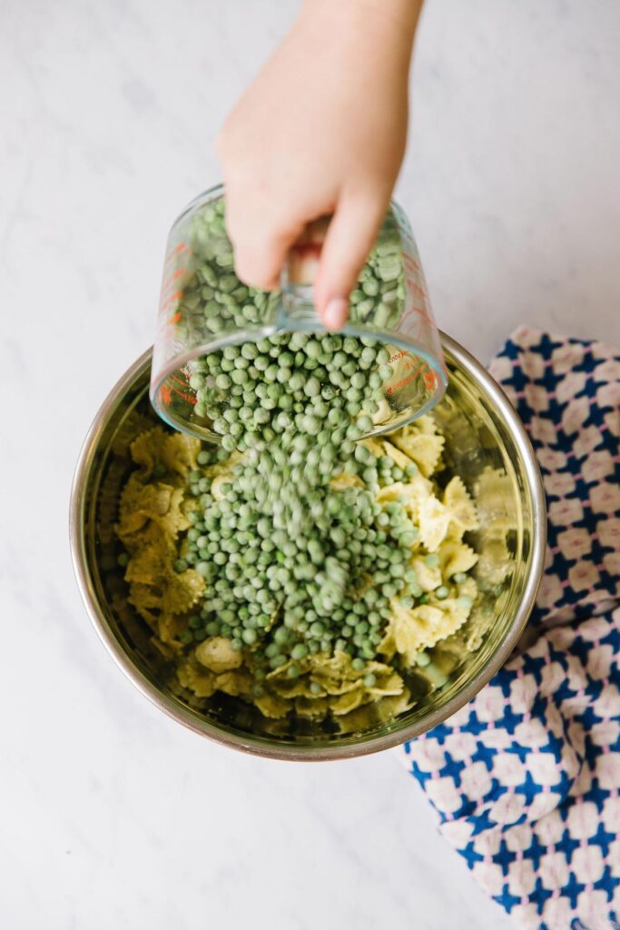 A silver bowl of pasta is on top of a pink and blue checked towel while a hand overhead pours frozen peas onto the pasta salad.