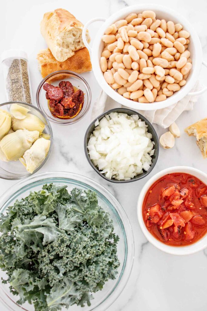 Ingredients to make a vegan freezer meal - bowl of white beans, onion, tomatoes, artichoke hearts, garlic, bread, spices and kale.