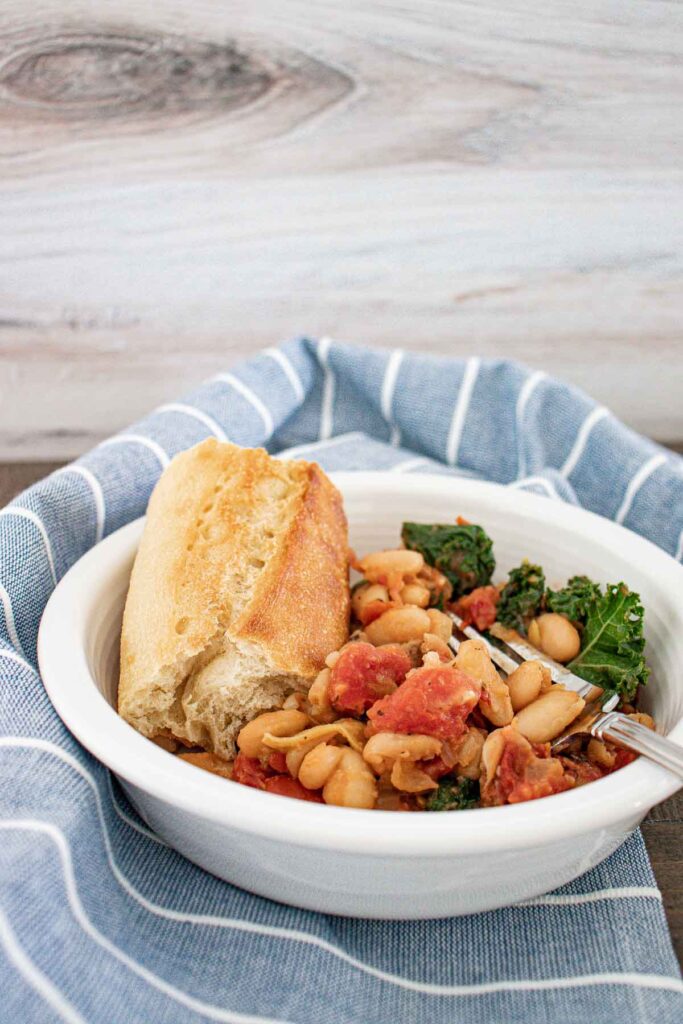 A blue and white striped towel with a white bowl of white bean and kale skillet meal with a piece of bread and a silver fork in it.
