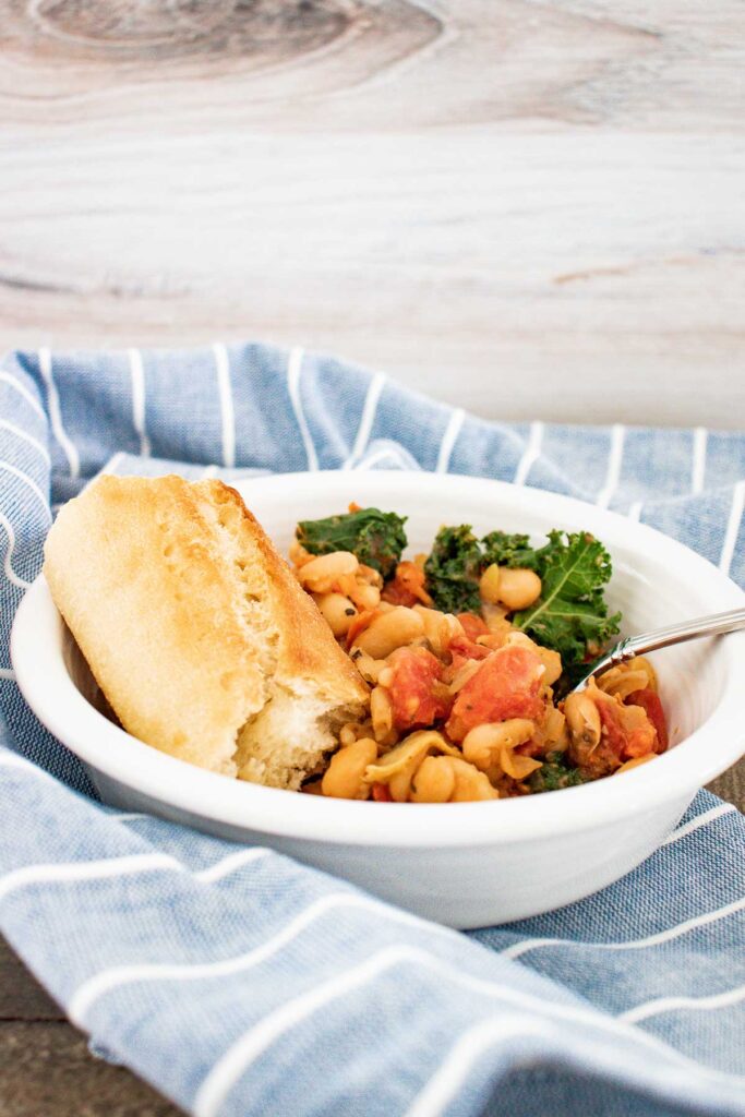 A blue and white striped towel with a white bowl of white bean and kale skillet meal with a piece of bread and a silver fork in it.