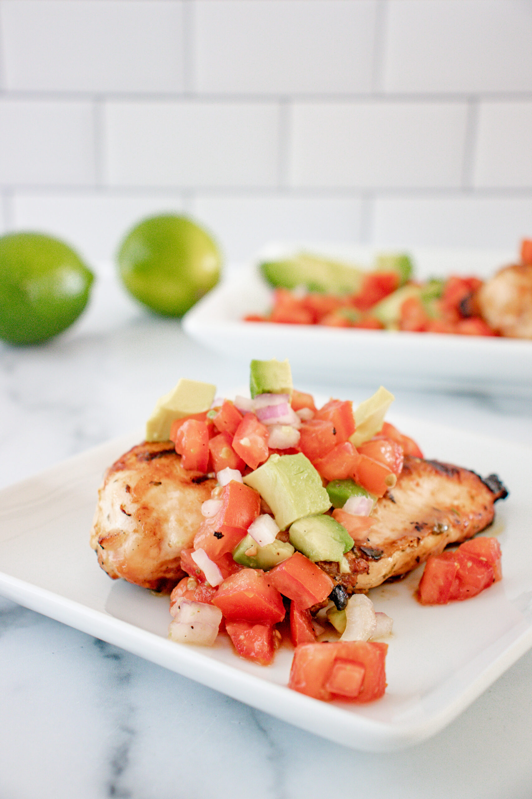 2 white dishes of lime cilantro chicken with avocado salsa and whole limes on the side.