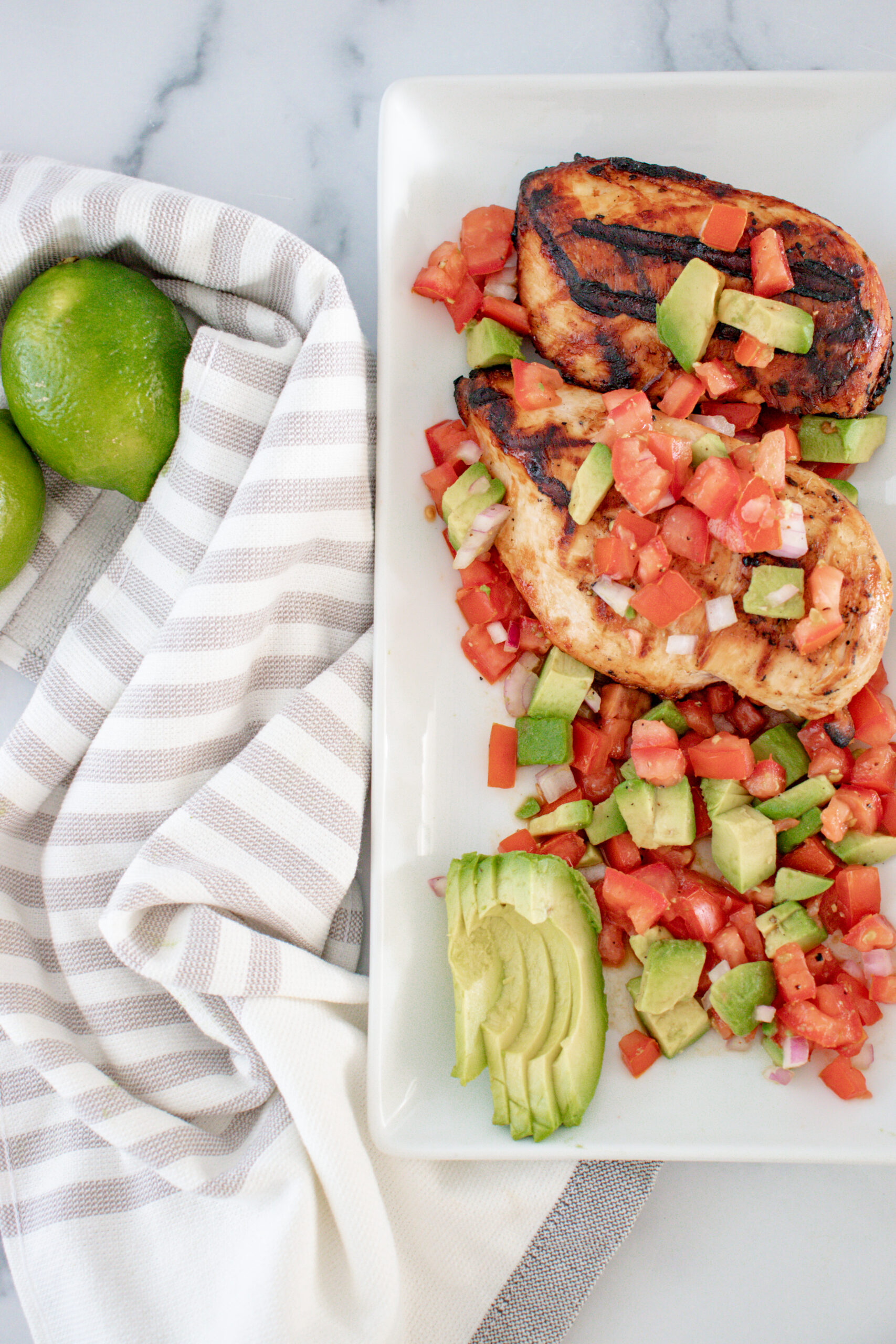 A white dish of lime cilantro chicken with avocado salsa with limes and a white dish cloth on the side.