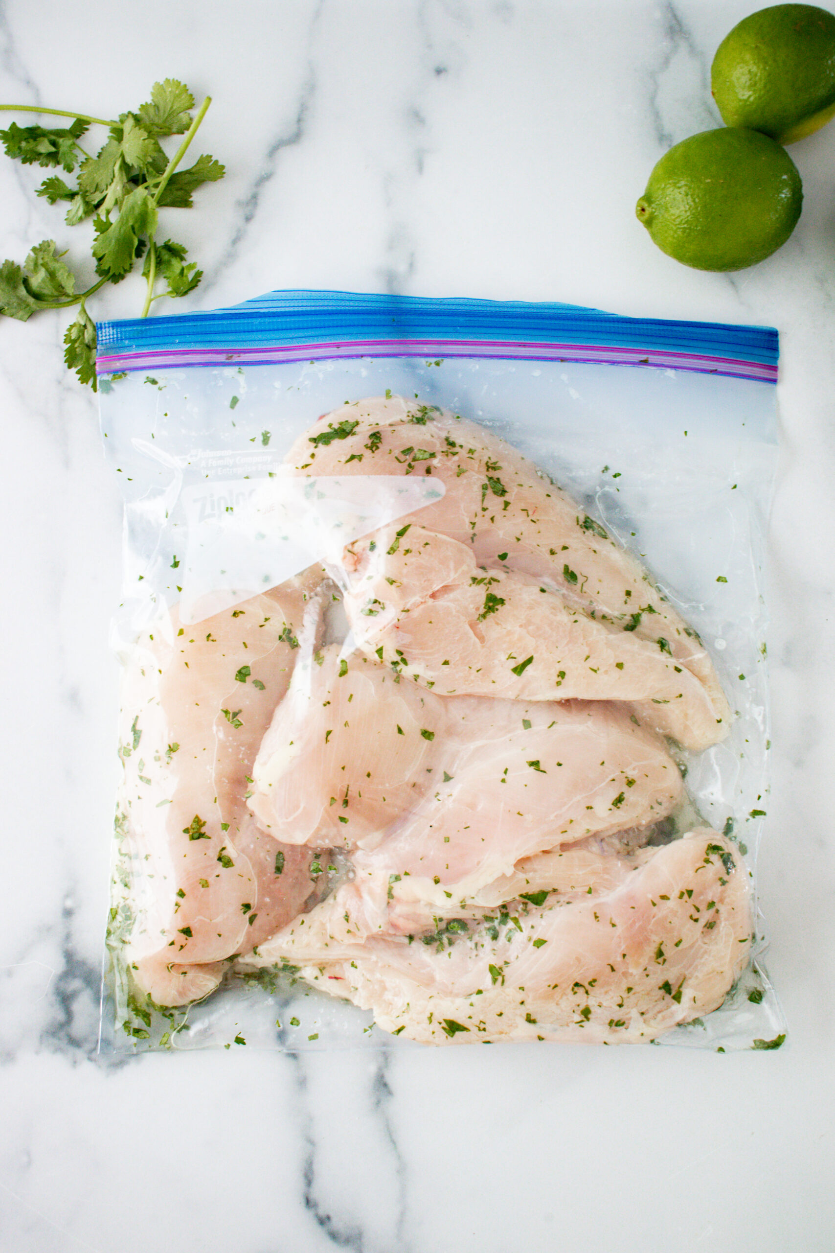 A resealable gallon bag with raw chicken breasts and seasonings with lime and cilantro on the side.