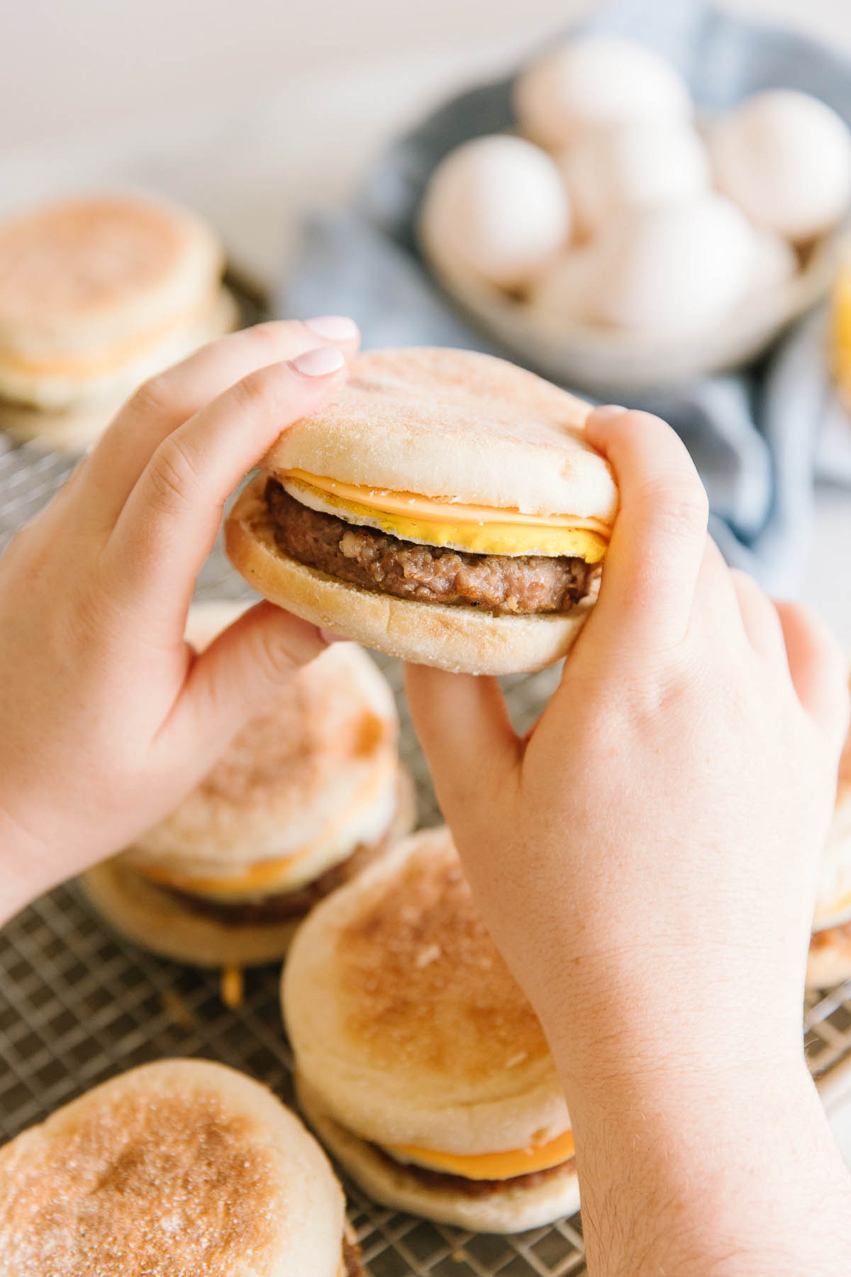 A pan with a wire tray with 6 breakfast sandwiches on it with  two hands holding one sandwich and a bowl of eggs on the side.
