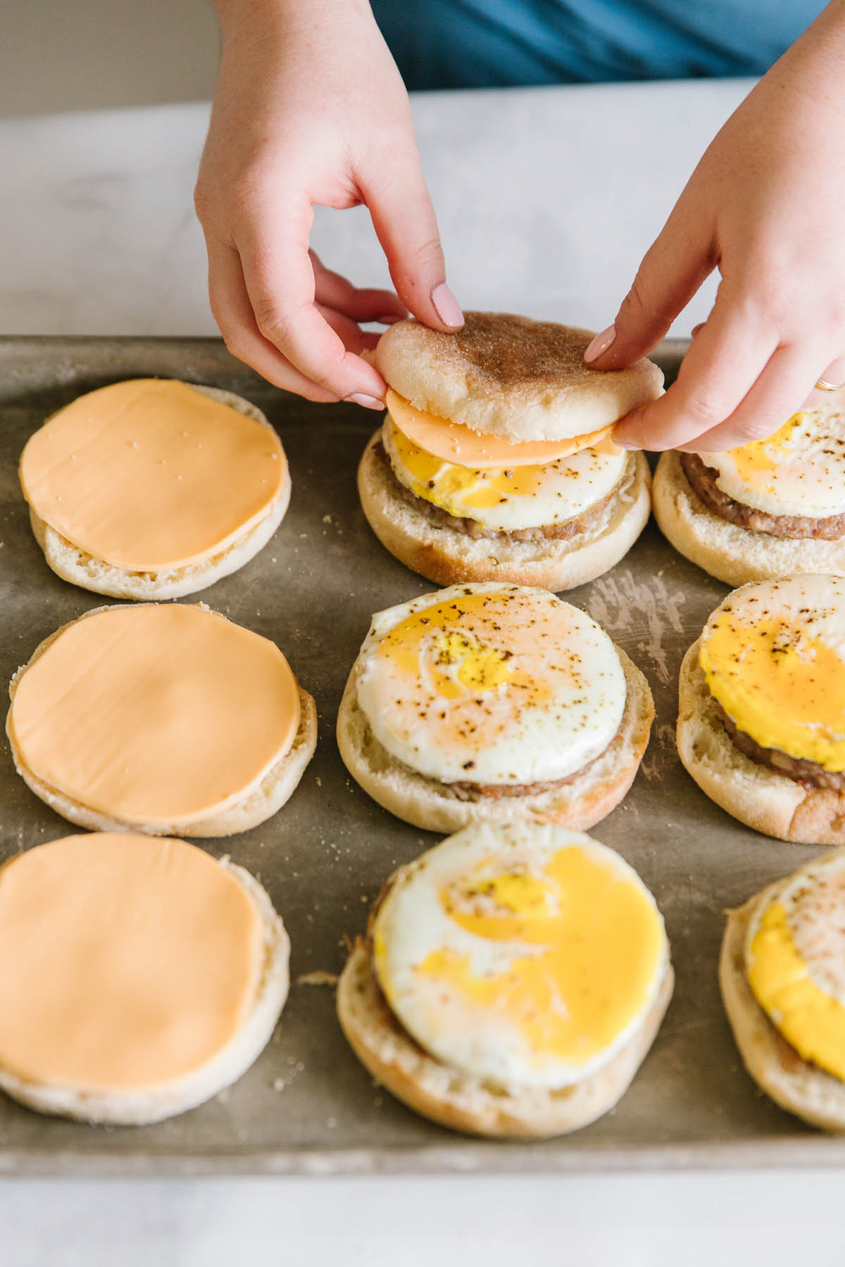 A pan with English muffins, cheese, eggs and sausage on them with two hands adding a top English muffin to one sandwich.