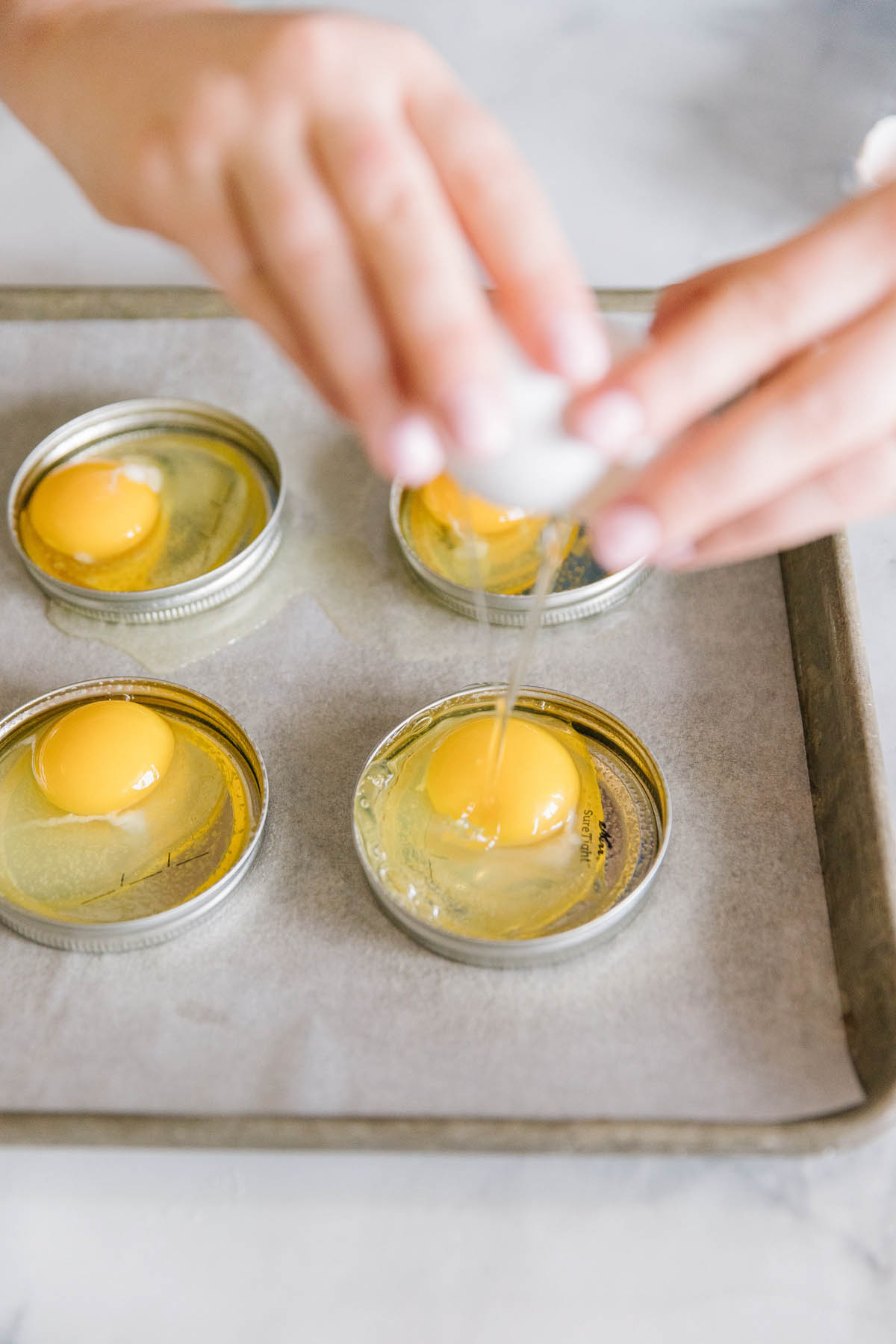 A pan with parchment paper with mason jar lids where a pair of hands have broken eggs into them.