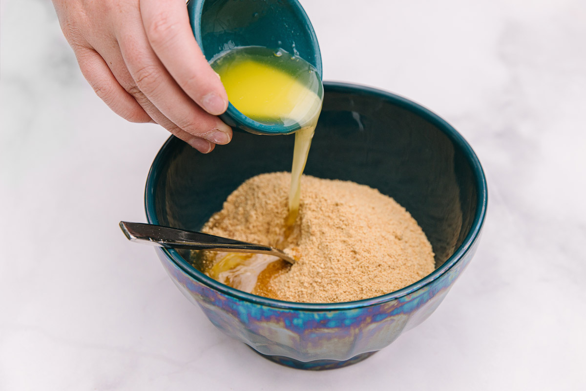 A bowl with graham crackers and brown sugar with a hand pouring a small bowl of butter into it.