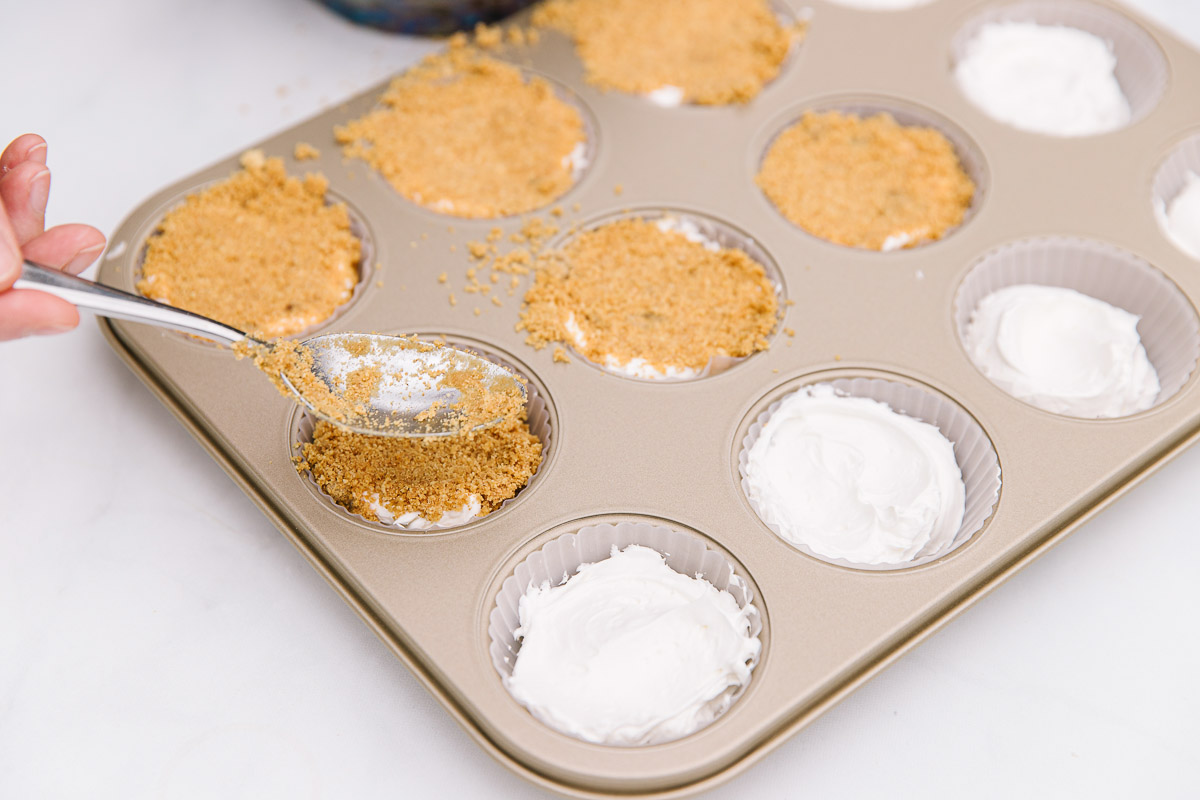 A cupcake pan with silicone liners with filling and a hand with a metal spoon putting graham cracker crumbs on top.