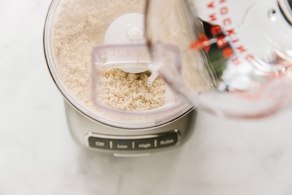 A food processor with dry ingredients with a measuring cup with water pouring in it.