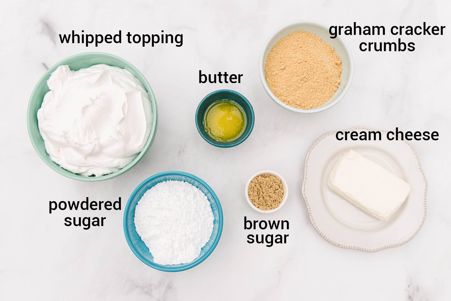 A white marble counter top with small bowls full of whipped topping, powdered sugar, brown sugar, butter, graham crackers, and a plate with cream cheese on it.