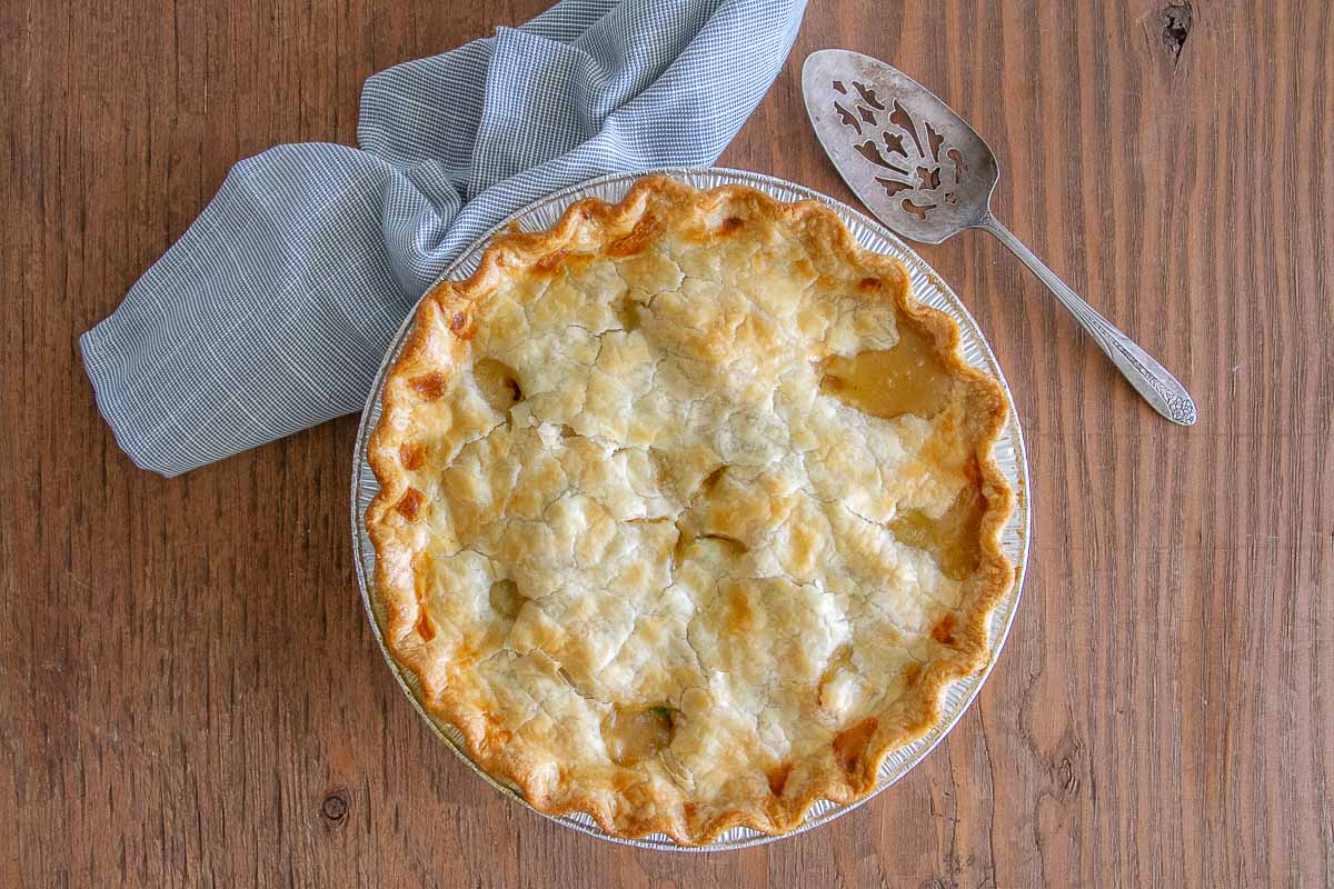 A whole chicken pot pie on a wooden background with a serving spoon and a white and blue towel.