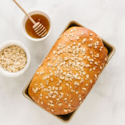 Loaf of honey oat bread in a bread pan with honey and oats.