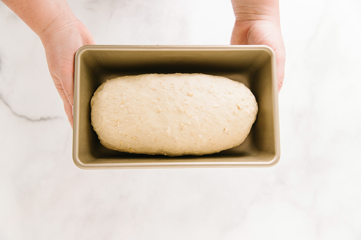 Two hands are holding a bread pan with a loaf of dough in it.