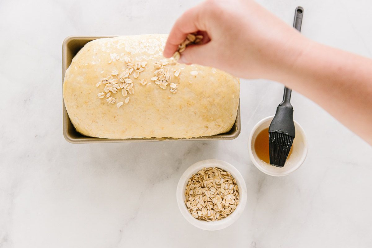A bread pan with risen dough in it with a hand sprinkling oatmeal on top. To the side is two bowls of honey and oatmeal.