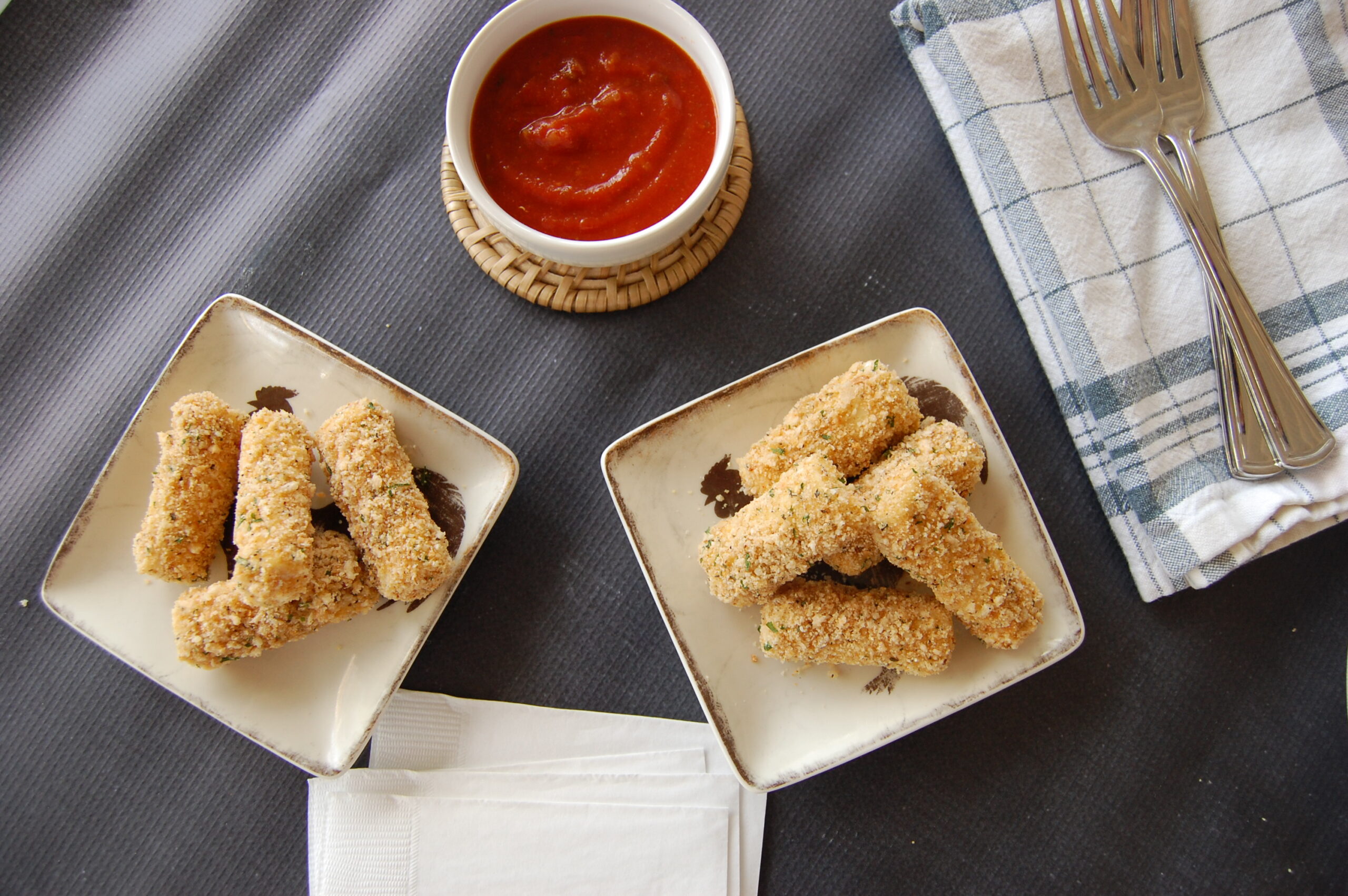 Two plates with mozzarella sticks piled on it with a jar of marinara sauce and a napkin with 2 forks on it.