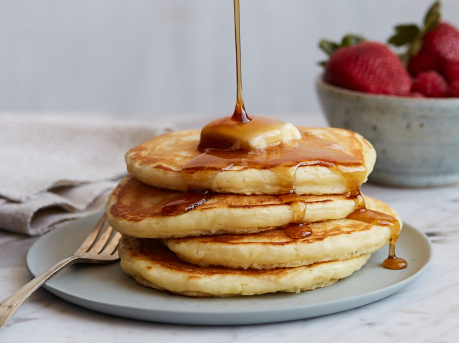A gray plate with a stack of four pancakes with a pat of button on it with syrup pouring down on it. There is a fork on the plate and a bowl of strawberries on the side.