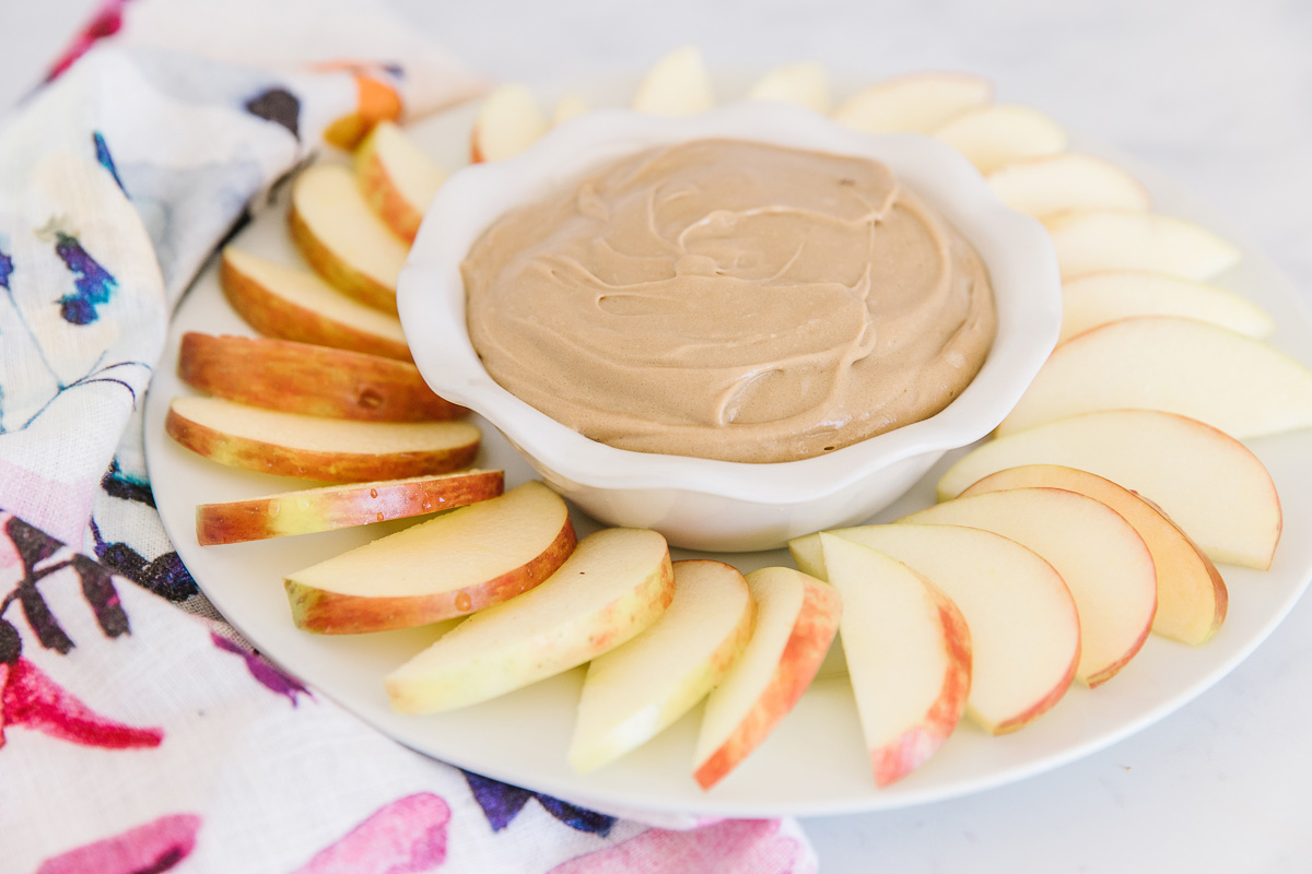 A white dish filled with apples with a bowl of brown apple dip with a flowered towel peeking from the side.