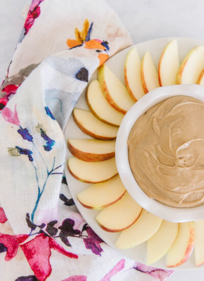 Tray of apple slices arranged around a bowl of cream cheese apple dip with a floral cloth napkin.