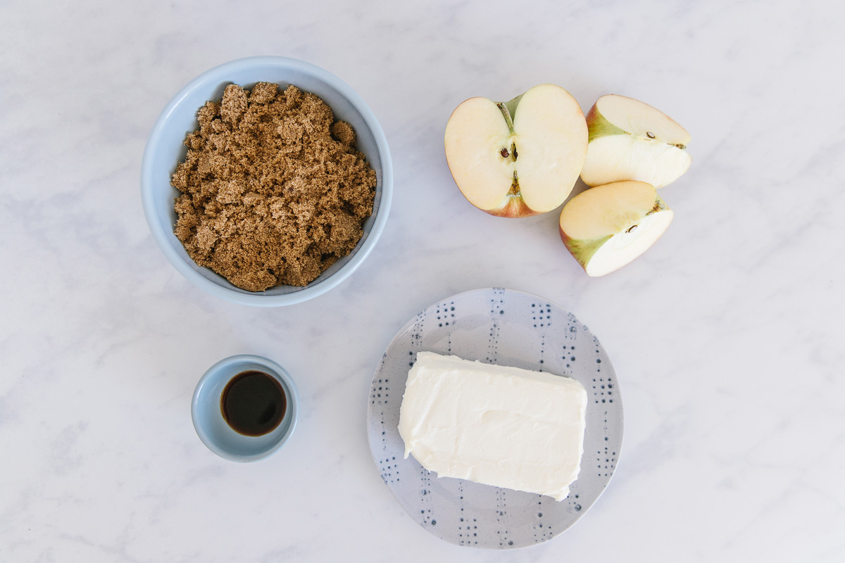 A blue  bowl of brown sugar, cut apples, a blue bowl of vanilla and a block of cream cheese.