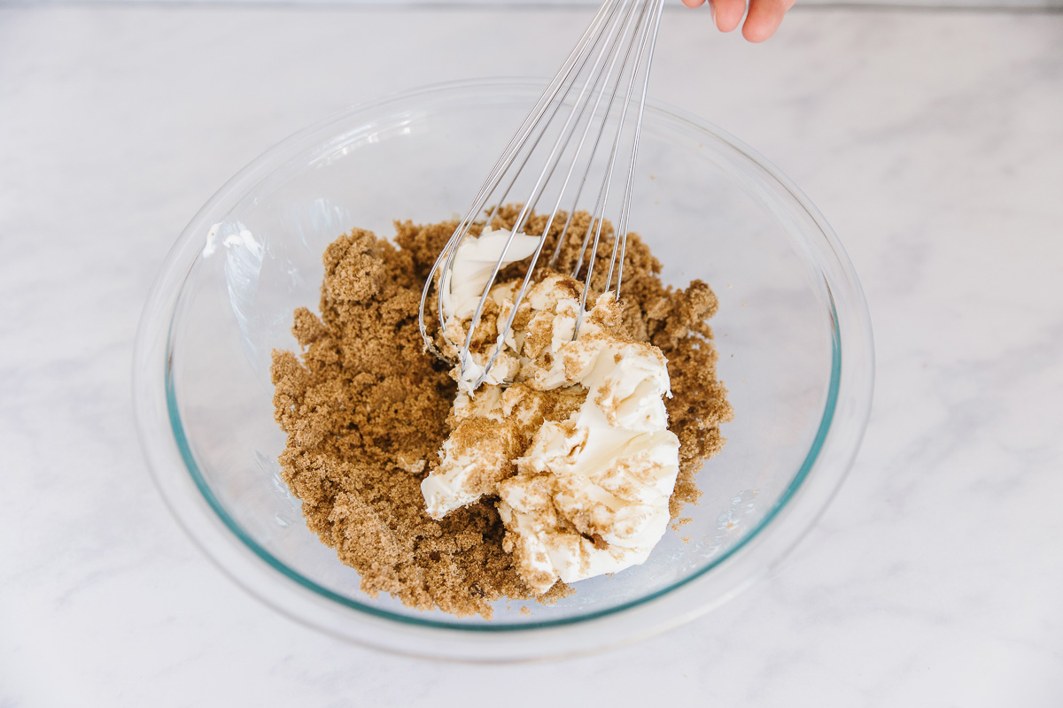 A glass bowl of brown sugar, vanilla, and cream cheese with a whisk mixing it.