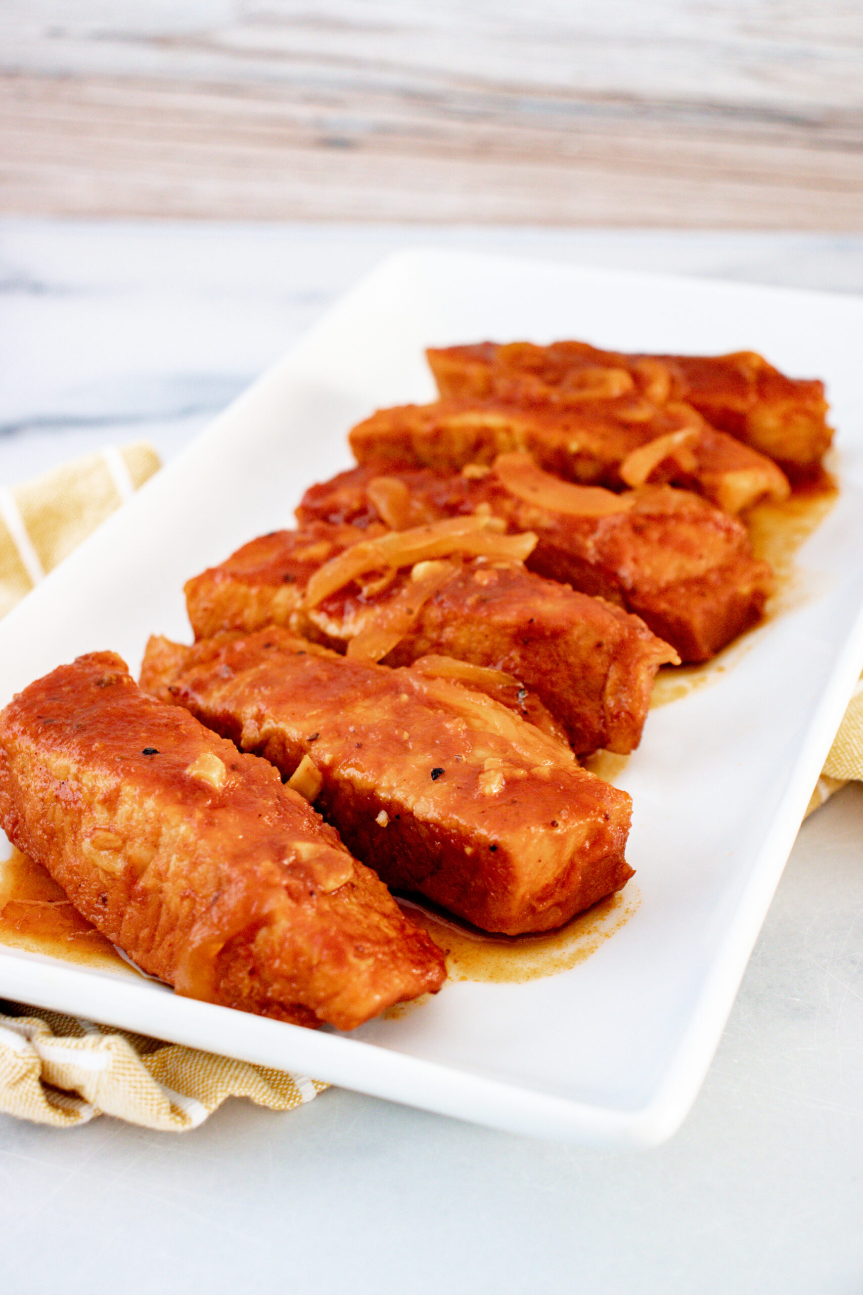 A rectangular white plate with cooked boneless ribs with sliced onions on it.