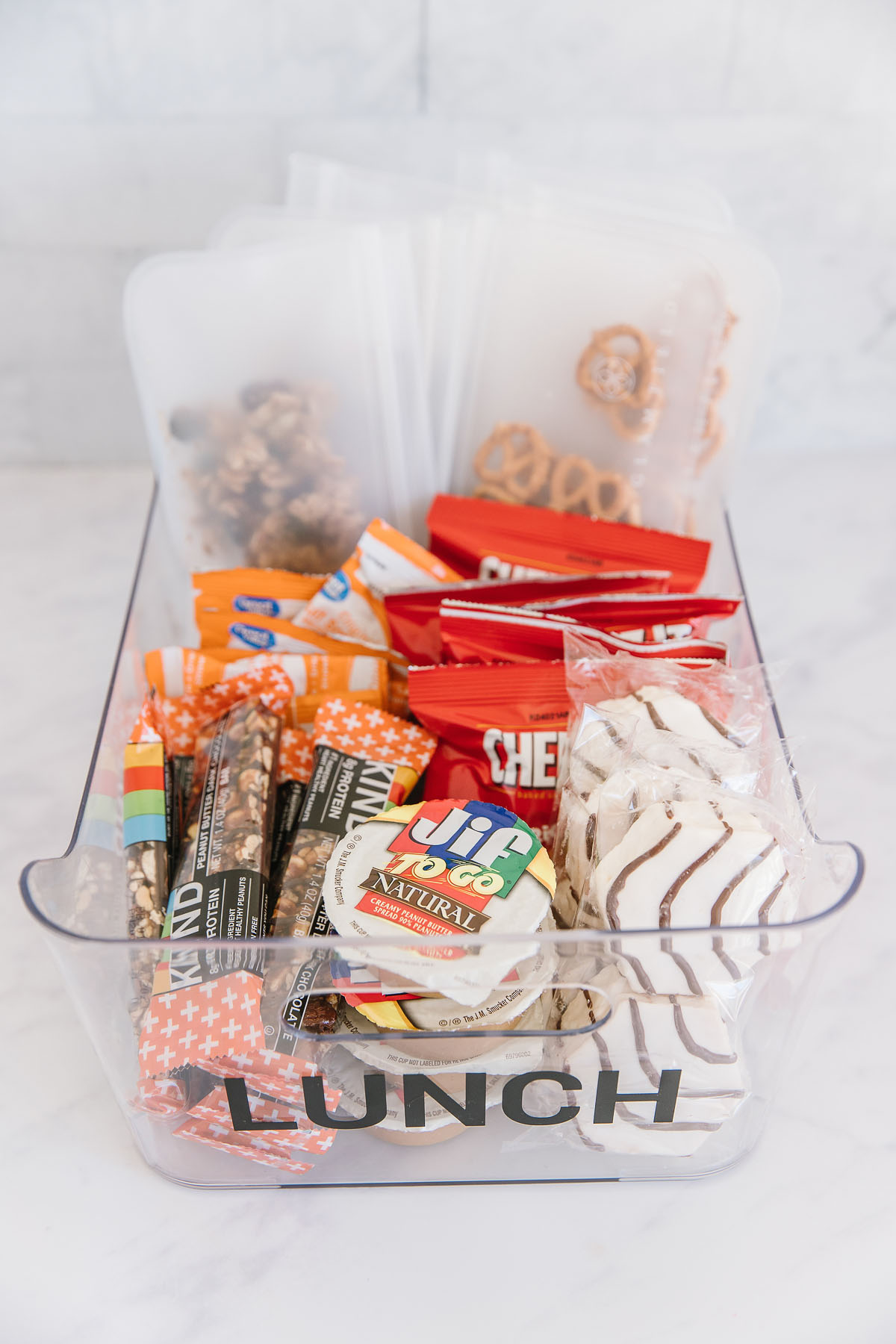 A plastic container with the word lunch on it filled with packaged snacks, nuts, peanut butter, crackers and pretzels. Perfect school lunch ideas!