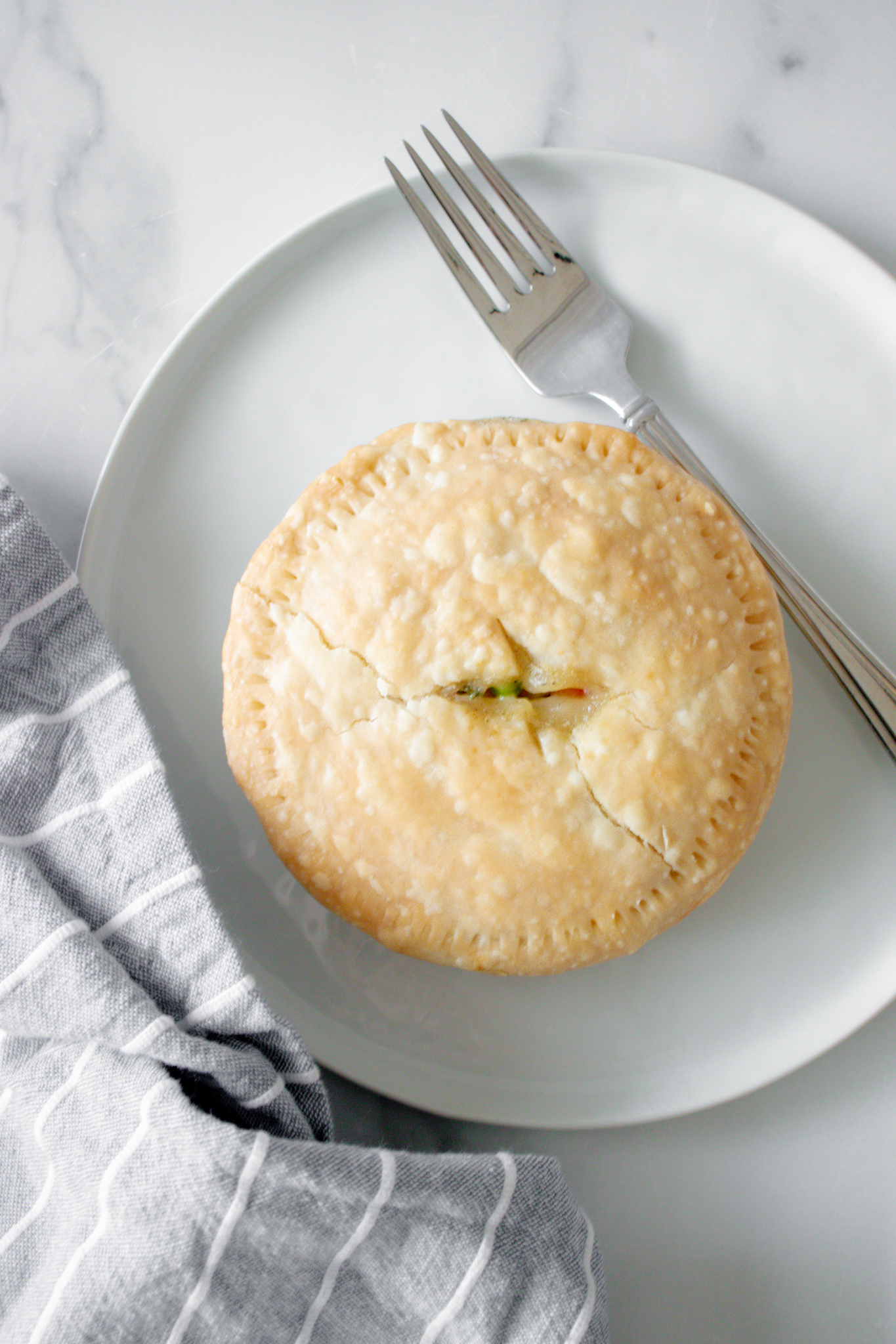 A white plate with a single chicken pot pie with a metal fork and a gray and white towel.