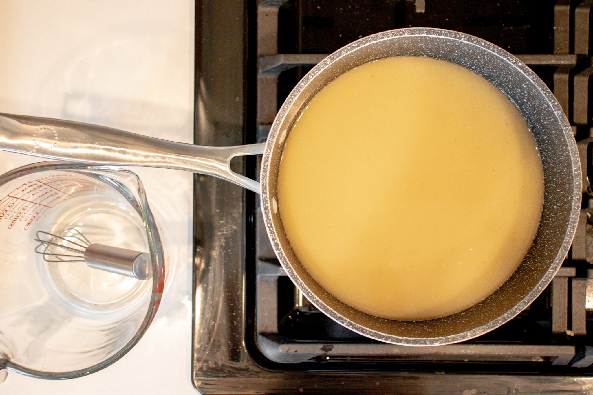 A stovetop with a metal saucepan with yellow liquid and a glass measuring cup with a little whisk on the side.   
