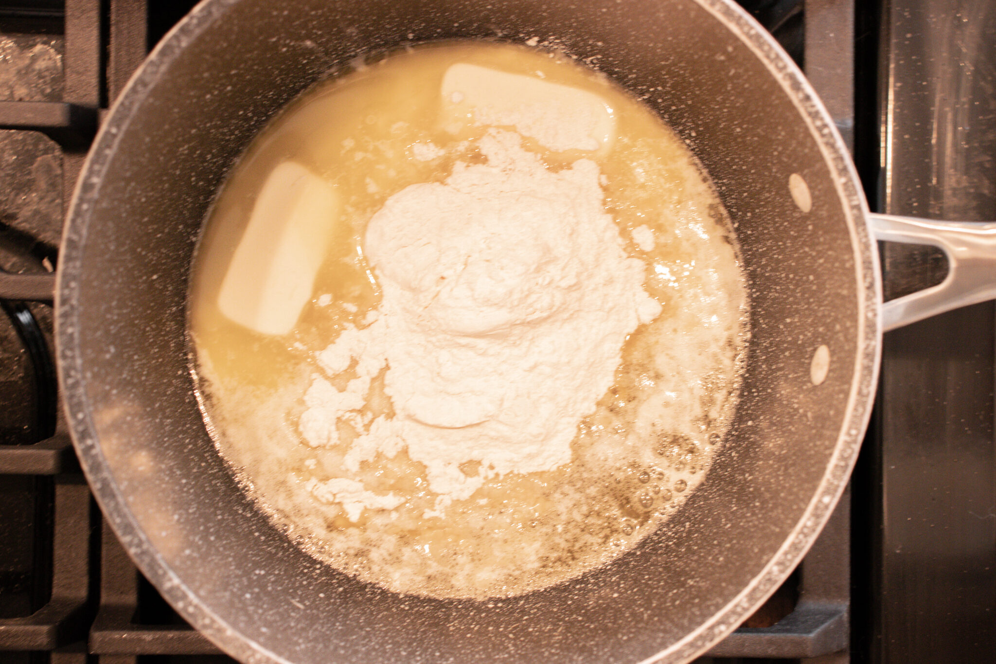 A metal saucepan on a stovetop with melting butter, flour and chicken stock.