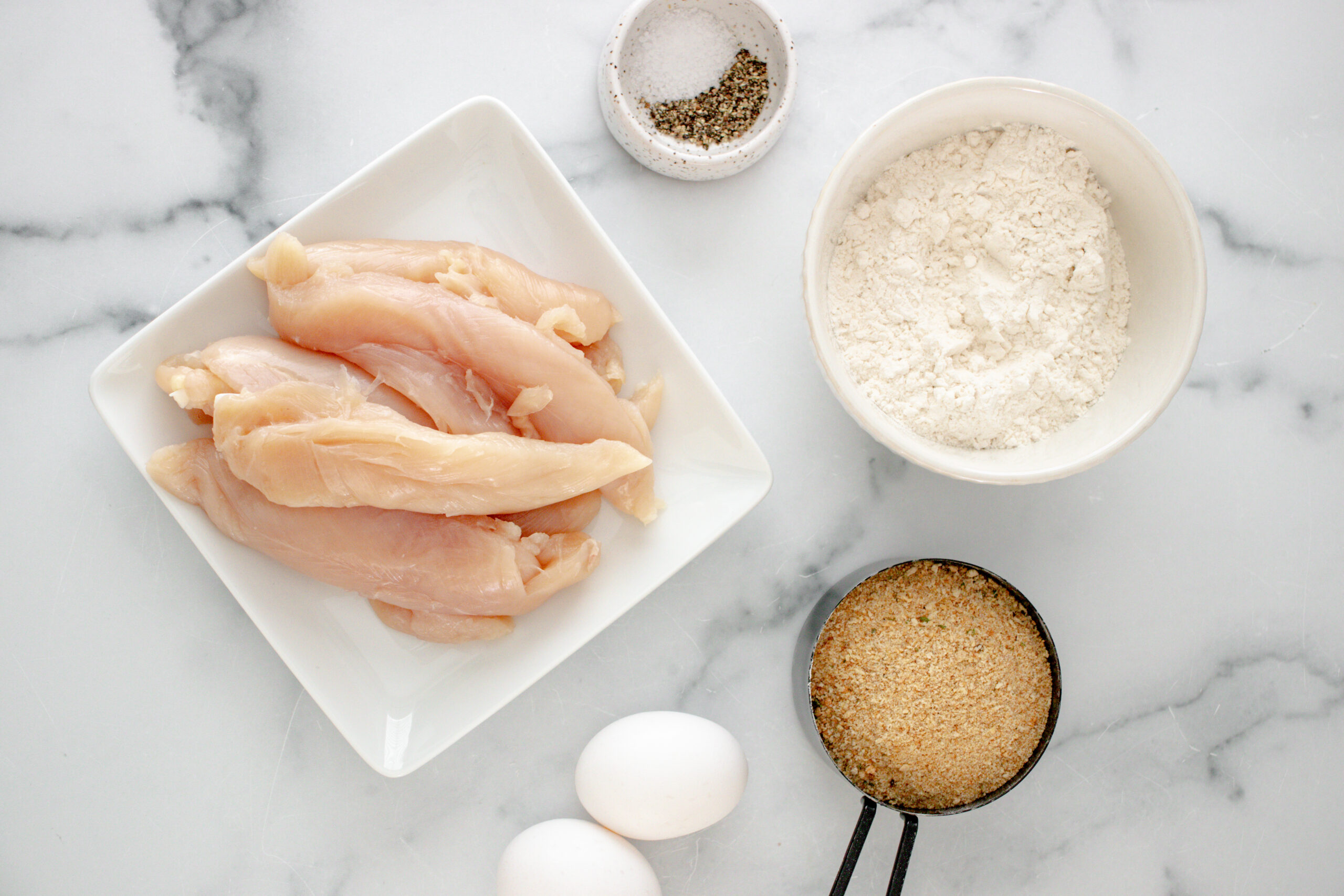A white plate with raw chicken and a white bowl of flour, eggs, panko and seasonings.