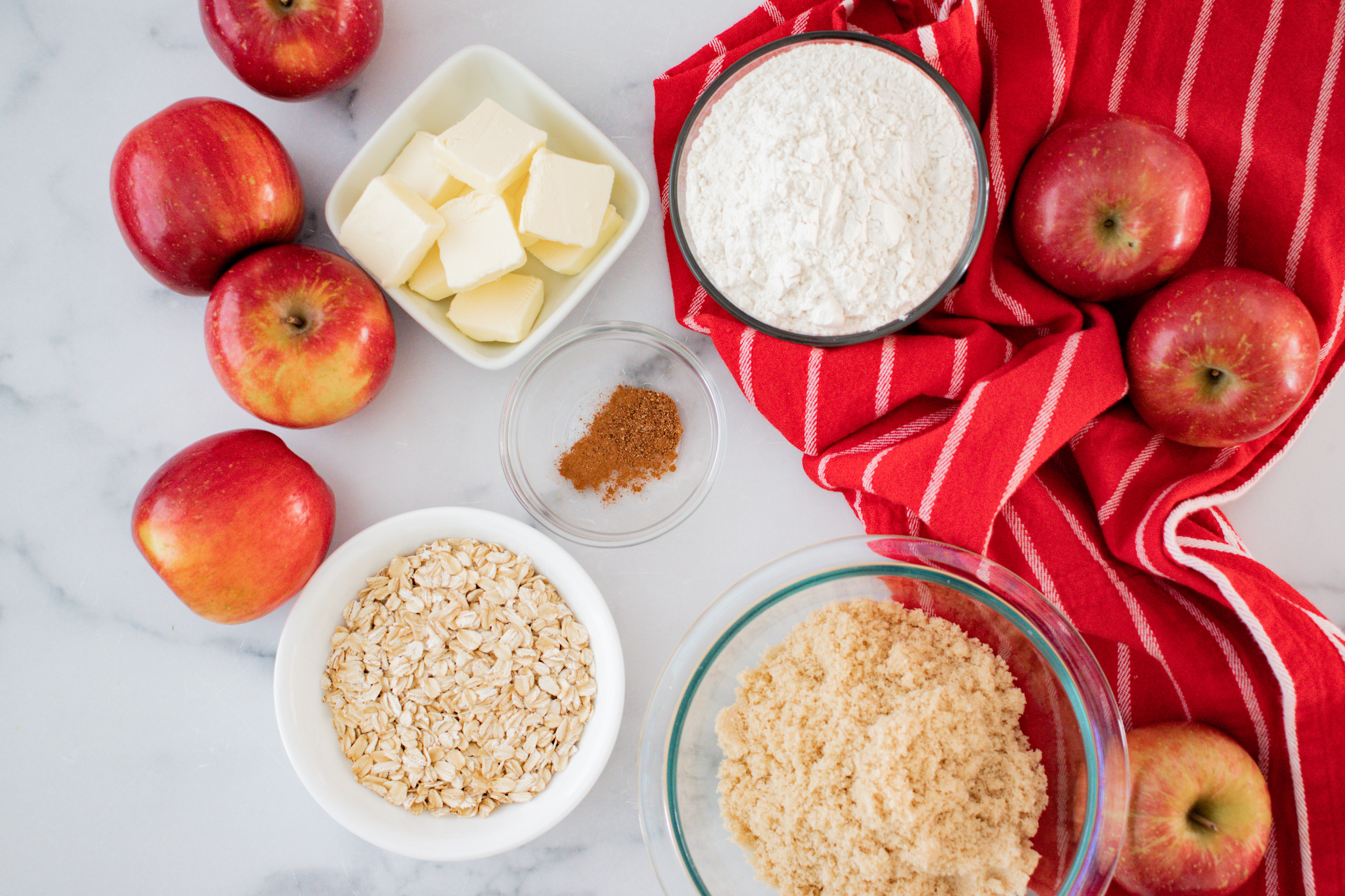 ingredients for apple crisp including butter in a white bowl, flour, cinnamon, oats and brown sugar in individual bowls and a red napkin.