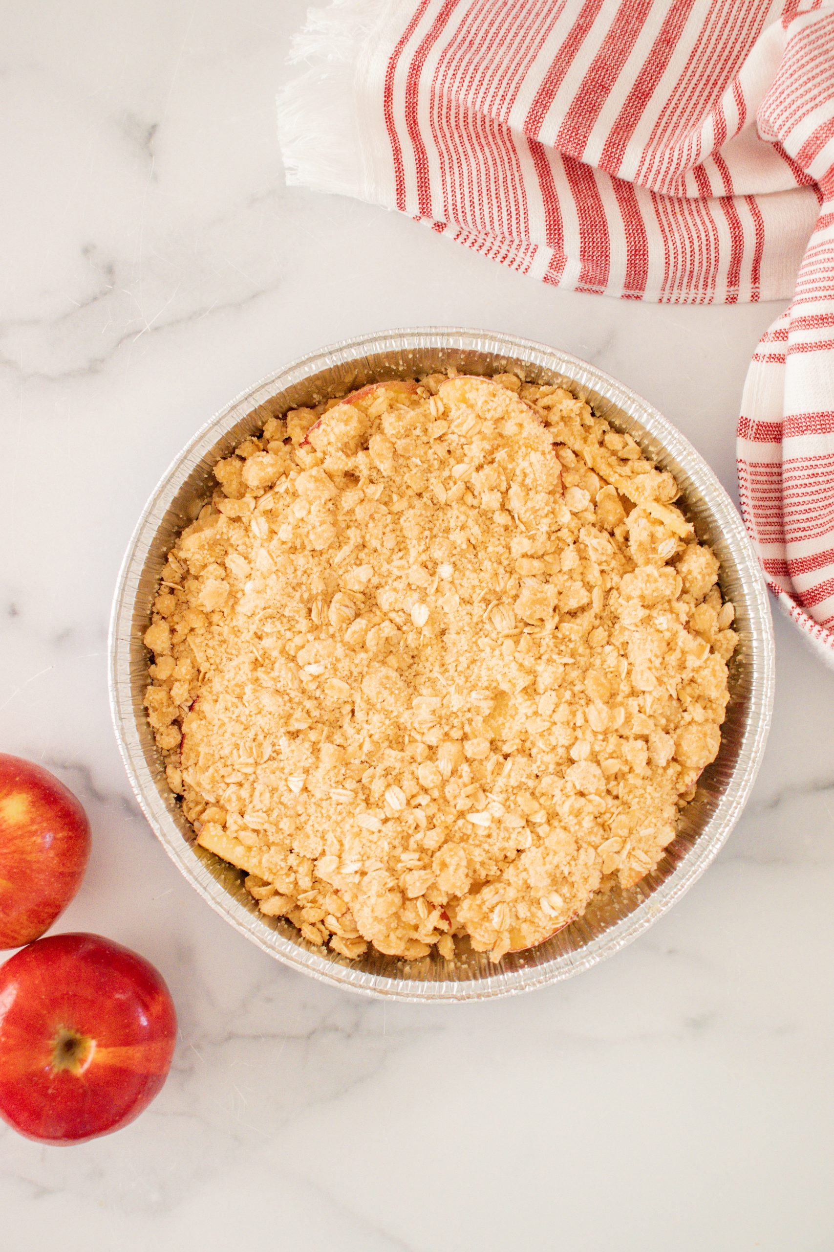 round foil pan with apple crisp inside it and a striped red and white towel in background and two red apples