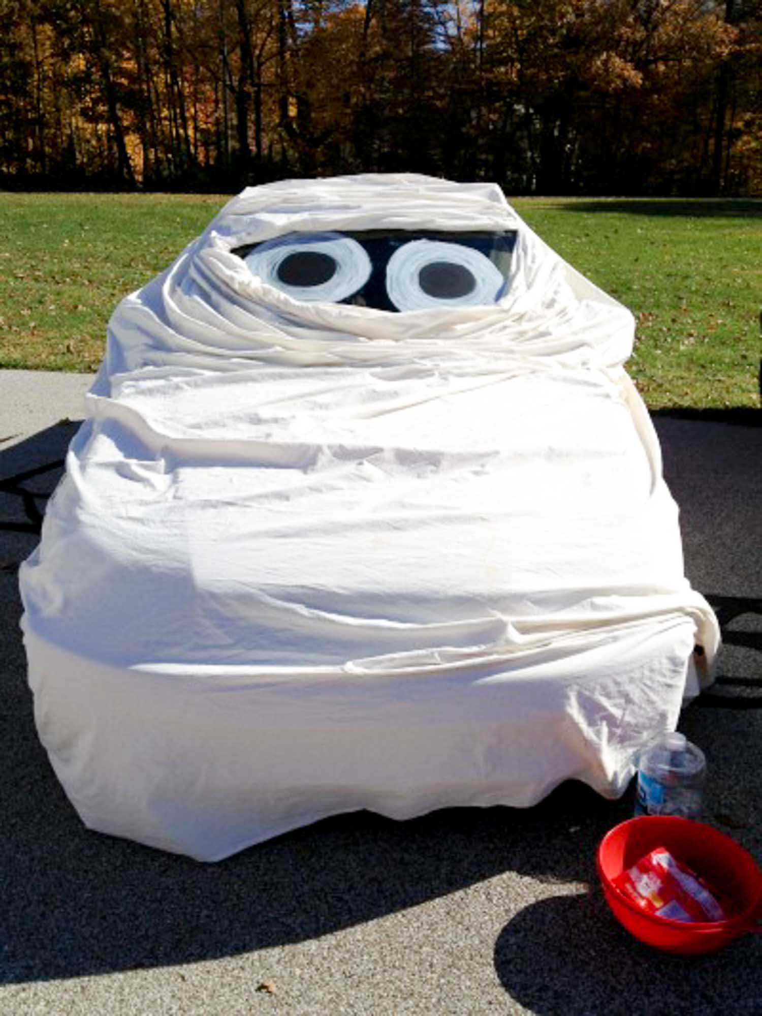 A car wrapped in a white sheet and eyes on the windshield with a bowl of candy next to it.